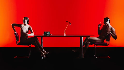 Two nude figures seated opposite each other at a long glass table against a vibrant red backdrop, styled by Alessandro Mensi.