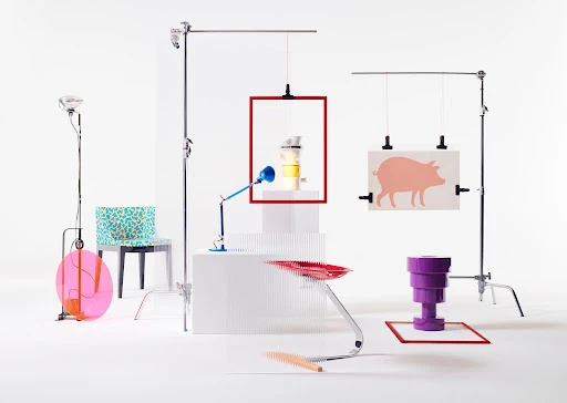 Studio still-life with a white backdrop, featuring a mix of furniture, lamps, and art objects in bold red, blue, and purple colors.