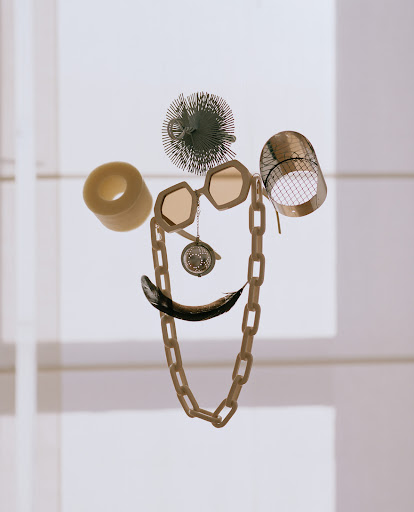 Still life of everyday objects arranged in the shape of a face, featuring a chain necklace, glasses, and black feather against a light background.