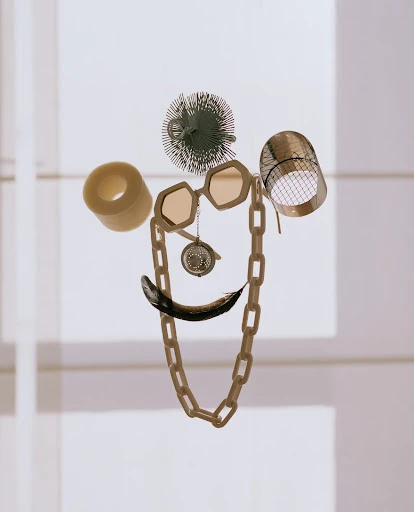 Still life of everyday objects arranged in the shape of a face, featuring a chain necklace, glasses, and black feather against a light background.