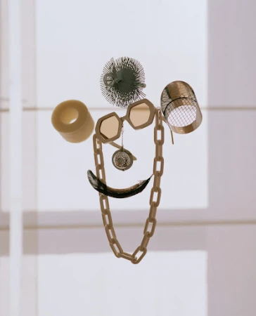 Still life of everyday objects arranged in the shape of a face, featuring a chain necklace, glasses, and black feather against a light background.
