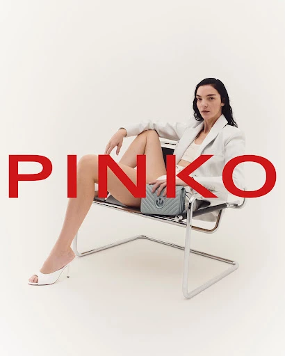 A stylish adult woman poses in a modern chair against a bright backdrop, showcasing a light blue purse and wearing a white blazer.
