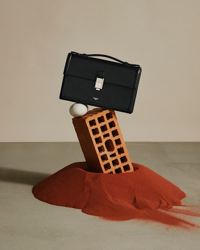 Black leather satchel resting on top of an egg and a brick, above a pile of red powder, all against a neutral studio background.