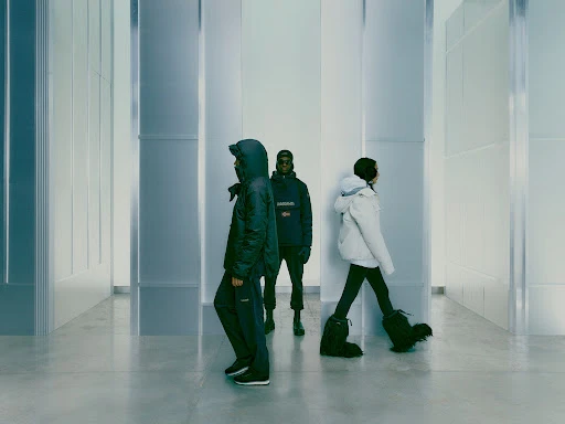 Three models in winter fashion walk through a modern corridor; one in dark blue hooded jacket and pants, one in white puffer jacket.