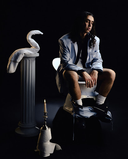 Person sitting on toilet, wearing suit, next to towel swan sculpture and lit candle against dark background, in a studio shot.