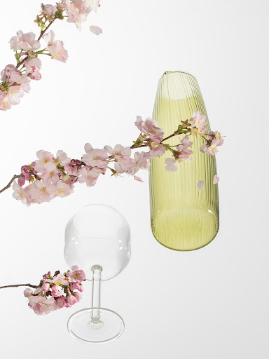 Still life of a clear wine glass, a chartreuse vase, and pink cherry blossoms on a white background creating a spring floral arrangement.