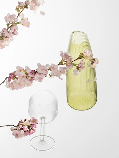 Still life of a clear wine glass, a chartreuse vase, and pink cherry blossoms on a white background creating a spring floral arrangement.