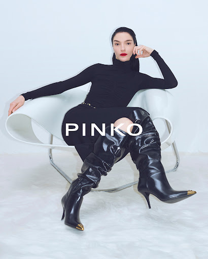 Elegant woman in black turtleneck, pants, and over-the-knee boots sits on a white chair in a light studio with visible brand logo.