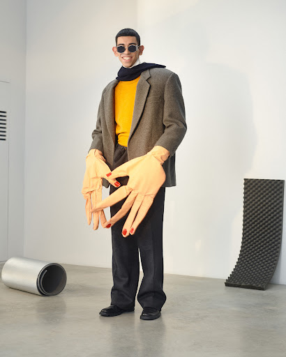 A man in a yellow sweater, jacket, and sunglasses smiles while wearing large, unrealistic hand-shaped gloves in a sparse photo studio.