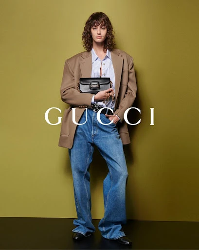 A woman in jeans, a blazer, and a button-up shirt poses with a Gucci purse against a yellow-green backdrop in a studio shot.