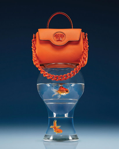 Bright orange Versace handbag with chunky chain, placed on a hourglass-shaped fishbowl with two goldfish, set against a blue gradient background.
