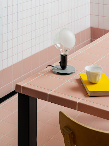 Close-up of a table with a desk lamp, book, and teacup, set in a room with tiled walls and floor, creating a minimalist, clean aesthetic.