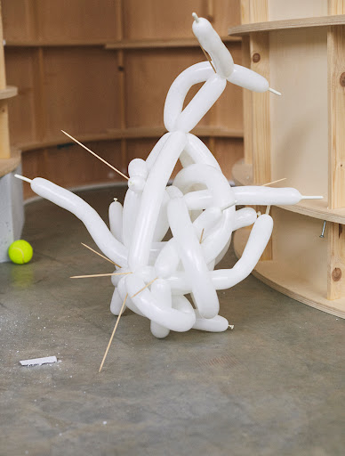 Sculptural arrangement of twisted white balloons and wooden skewers against a partially constructed wood background. Bright, whimsical.