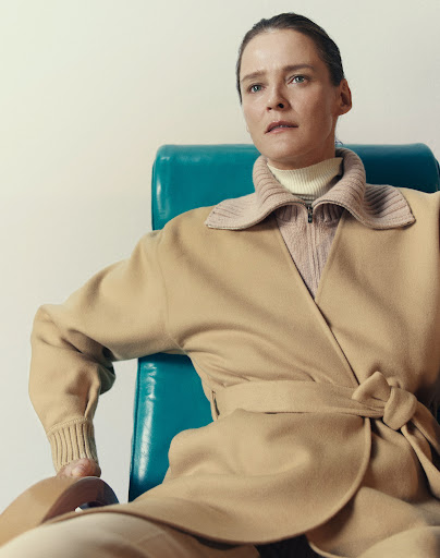 Portrait of a fair-skinned adult female seated on a teal chair, wearing a beige belted coat, in a brightly lit indoor setting.