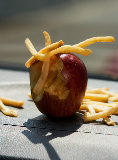 Close-up of a bitten red apple with french fries inserted into the missing section, resting on a gray surface with fries scattered around it.