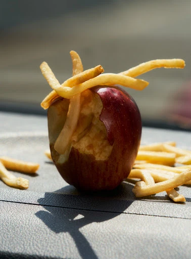 Close-up of a bitten red apple with french fries inserted into the missing section, resting on a gray surface with fries scattered around it.