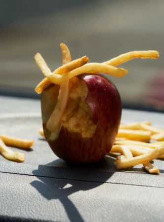 Close-up of a bitten red apple with french fries inserted into the missing section, resting on a gray surface with fries scattered around it.