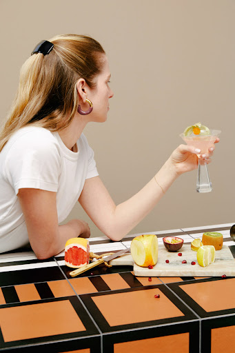 Woman in white t-shirt holds pink drink, sitting at orange and black tile table with fruit, and marble cutting board indoors.