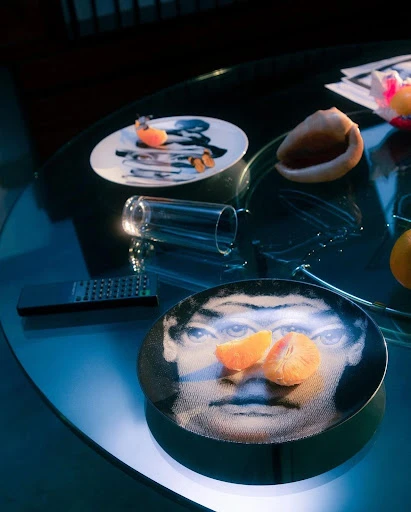 Low-angle indoor shot of a glass-top table featuring decorative plates, a glass, fruit, a seashell, and a remote control in a dimly lit setting.