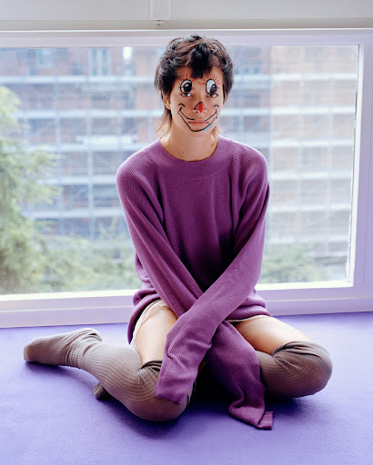 Woman with clown face paint sits in front of window, wearing a purple sweater and thigh-high socks, neutral facial expression, indoor setting.
