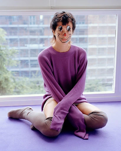 Woman with clown face paint sits in front of window, wearing a purple sweater and thigh-high socks, neutral facial expression, indoor setting.