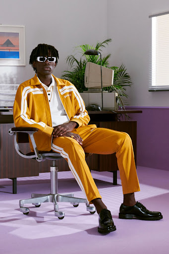 Man in yellow tracksuit and sunglasses poses in office chair near desk with vintage computer and indoor plant in light purple room.