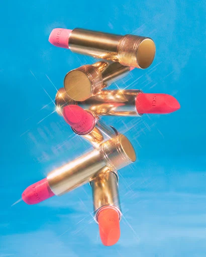 Tower of coral lipsticks with gold tubes stacked against a vibrant blue background, beauty and fashion product shot.