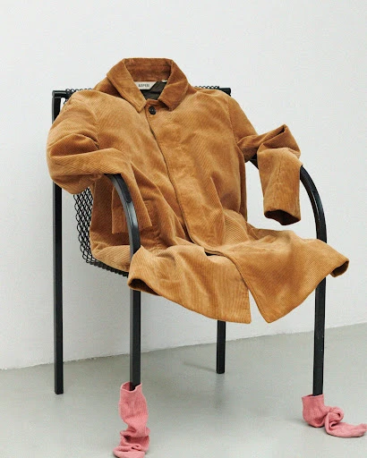 Corduroy coat draped on black metal chair with pink socks on chair legs against a minimalist white background in a photo studio.