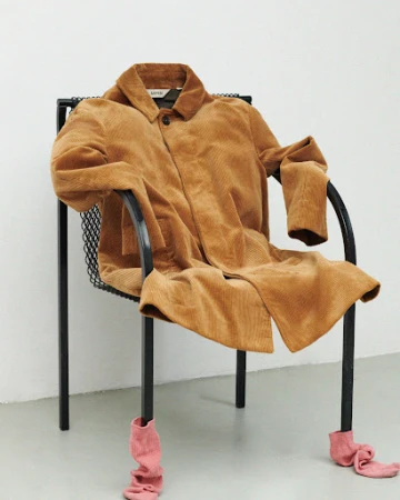 Corduroy coat draped on black metal chair with pink socks on chair legs against a minimalist white background in a photo studio.