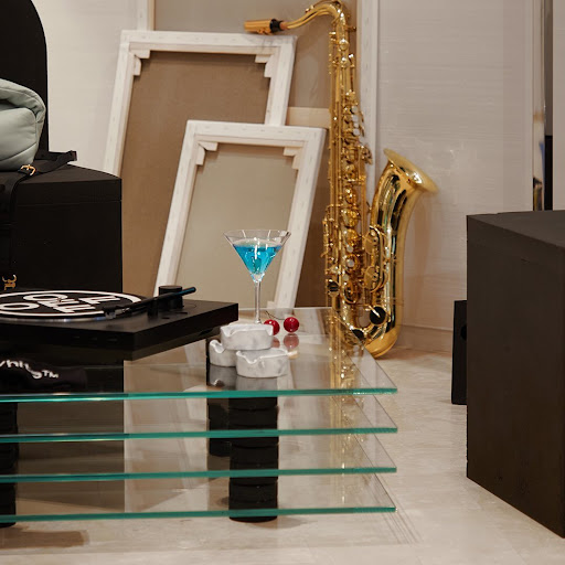 Saxophone and blue cocktail sit on a glass table with a record player and cherry garnish in a minimalist, neutral setting.