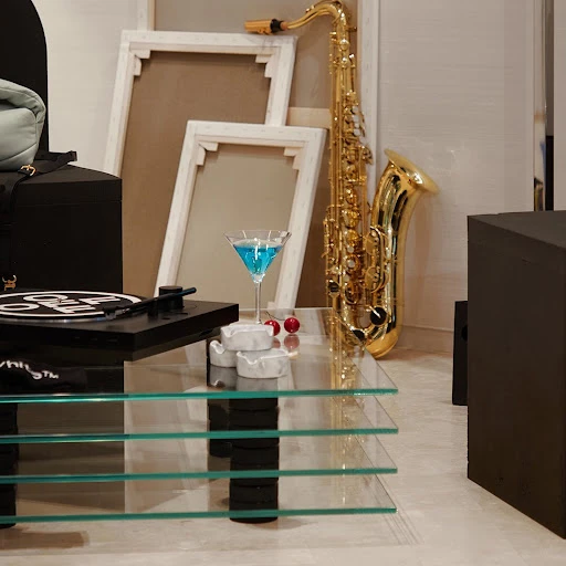 Saxophone and blue cocktail sit on a glass table with a record player and cherry garnish in a minimalist, neutral setting.
