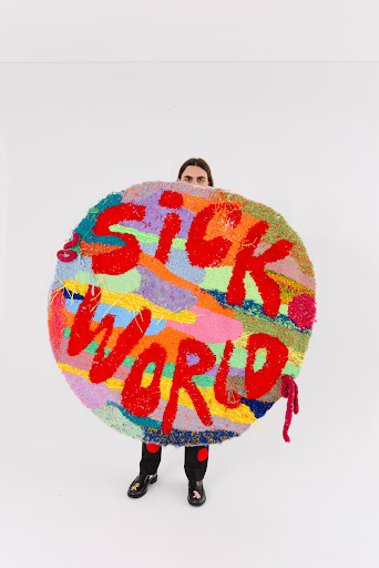 Person standing indoors holding a vibrant round rug with 'Sick World' text, featuring colorful yarns and abstract patterns on a white background.