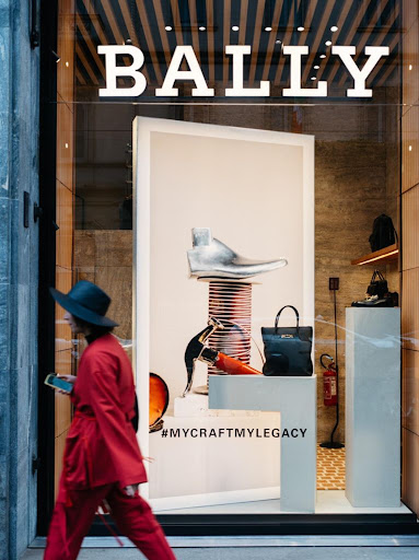 Woman in red walking past a Bally storefront with shoe sculpture, purses, and #MYCRAFTMYLEGACY campaign text on white display stands.