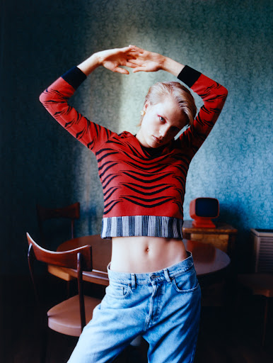 Indoor portrait of a slender woman in a red tiger-striped top and jeans, posing with her arms raised in front of a textured blue wall.