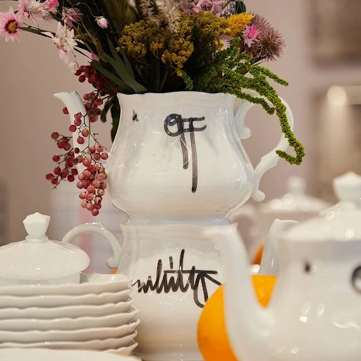 White ceramic teapots stacked with colorful flowers, orange, and white plates creating a bright, artistic still-life composition indoors.