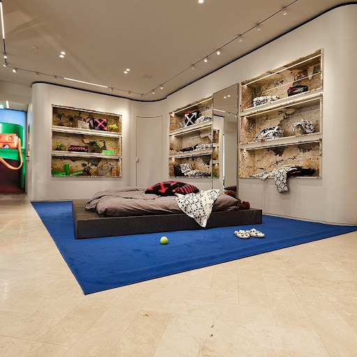 Modern bedroom display inside clothing store with bed on blue rug, stone-lined shelves showcasing clothes and accessories, and travertine flooring.