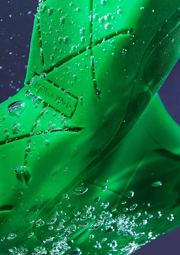 Close-up of a vibrant green Bottega Veneta rubber shoe with water droplets, set against a blurred, dark background.