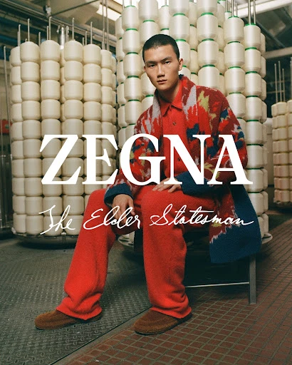 Asian male model in red suit and patterned jacket poses in industrial setting with stacked spools behind him, promoting Zegna menswear.