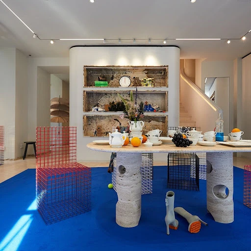 Interior of a store with a table set with dishes and fruit, surrounded by wireframe chairs on a blue floor, displaying modern design.