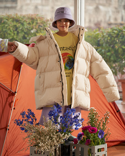 A young child wears an oversized cream puffer jacket and bucket hat. Flowers sit in the foreground; an orange tent in the background.
