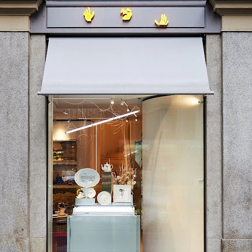 Storefront view with a gray awning, yellow hand-shaped signs, and a display of plates and teacups in the window.