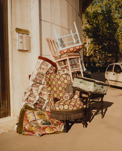 A stack of Gucci-patterned chairs, a footstool, and a folded quilt sit on a sidewalk next to a beige building and a white car on the street.