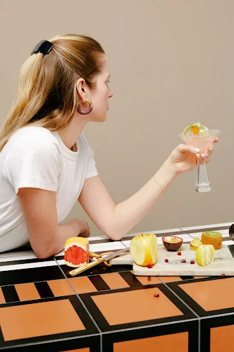 Woman holding a pink cocktail in profile view at a table with sliced fruits and geometric terracotta tiling