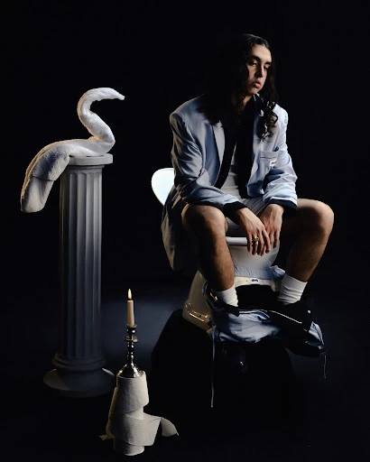 Man with long dark hair wearing a light blue blazer seated on a white pedestal with a classical column and a lit candle.