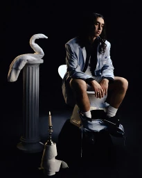 Man with long dark hair wearing a light blue blazer seated on a white pedestal with a classical column and a lit candle.