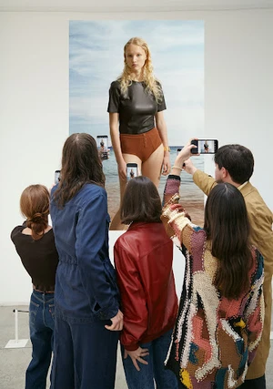 Group of people holding up smartphones to photograph a large portrait print of a blonde model on a white wall.