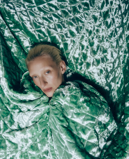 Adult fashion model enveloped in quilted light green fabric, portrait-style, indoor studio shot, featuring pale skin, blonde hair.