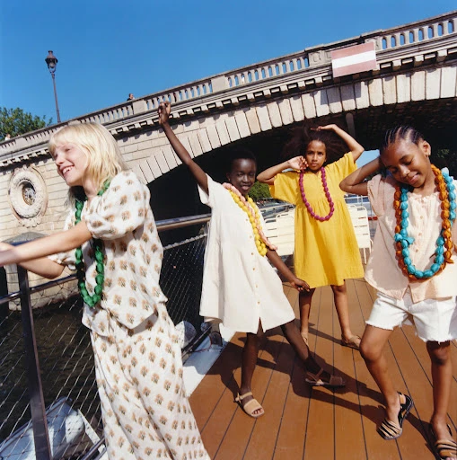 Four smiling children in summery outfits and colorful necklaces pose on a boat under a bridge, enjoying a sunny day in an urban setting.