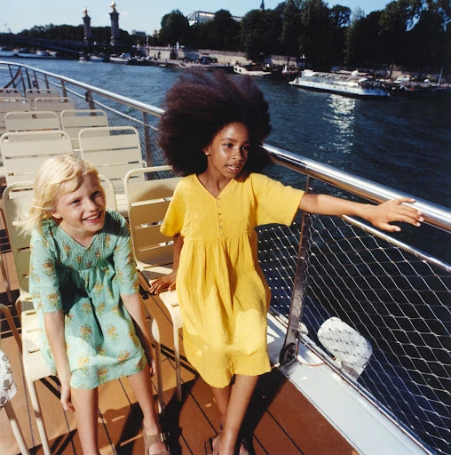 Two young girls in summer dresses sitting on a boat with a river, bridge, and architecture in the background on a sunny day.