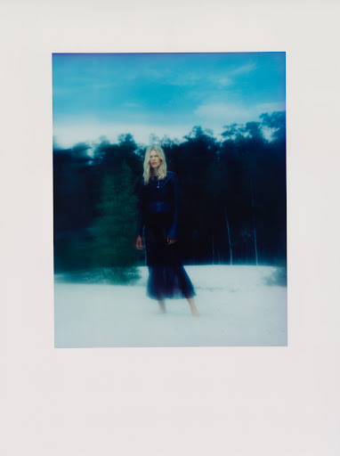 Blonde woman in a dark dress stands barefoot in a blurry, dreamlike outdoor scene with a forest and sky background in soft focus.